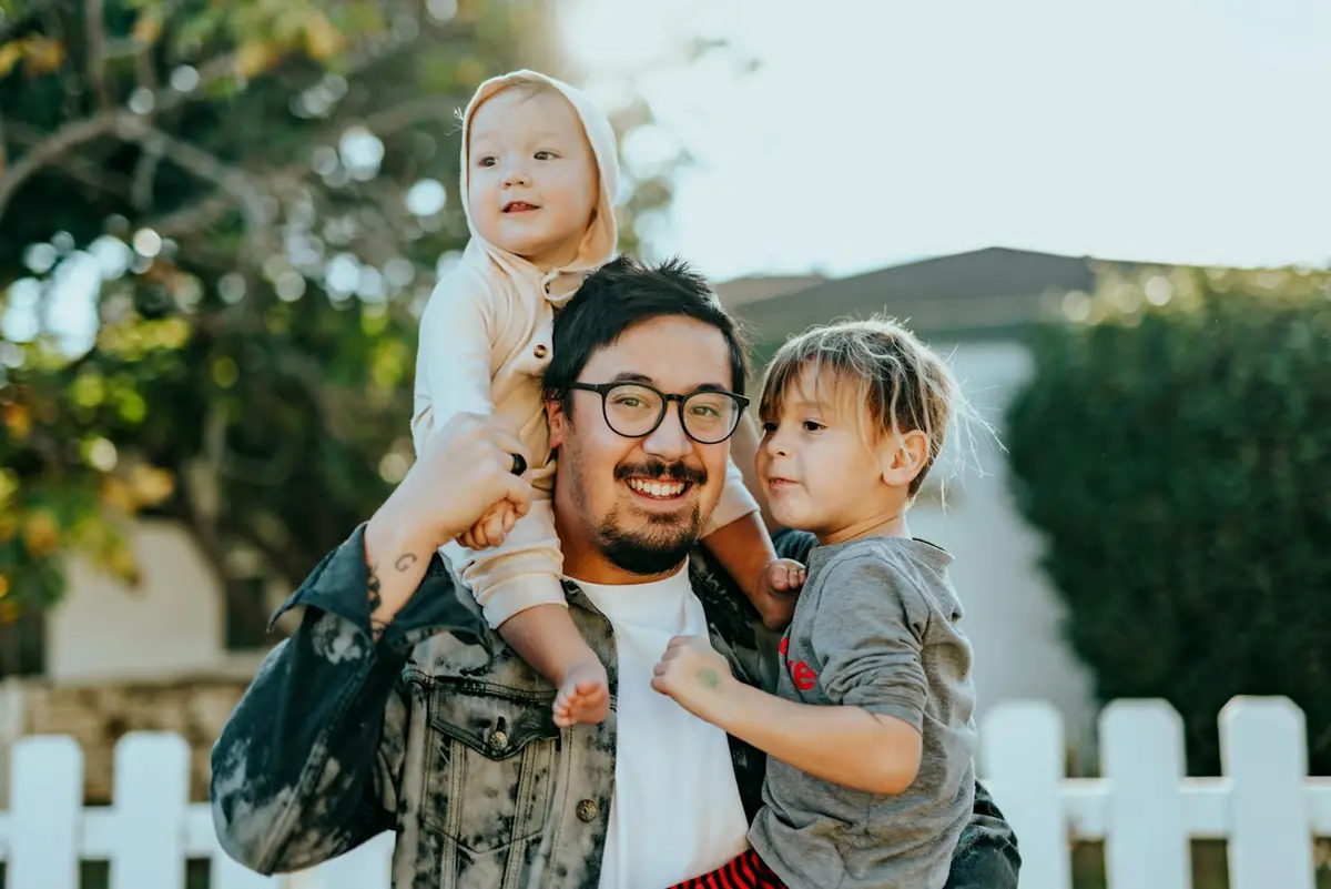 Parent carrying two young children in front of their home